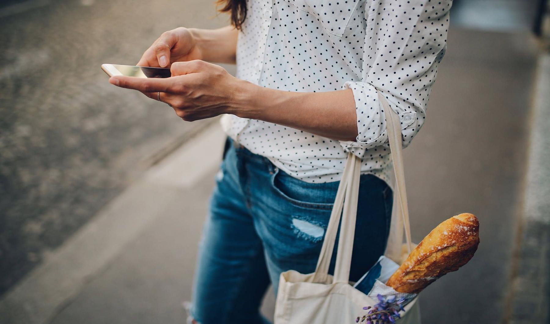 Close to the center of everything woman walks down the street and texts with a bag of groceries on her arm